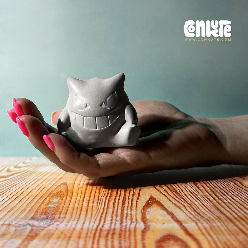 May include: A gray ceramic figurine of a ghost Pokemon character with a wide grin and large eyes. The figurine is sitting on a person's hand.