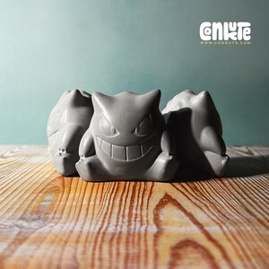 May include: Three gray ceramic figurines of the Pokemon Ghost-type, Gengar. The Gengar in the center is facing forward with a wide grin and large eyes. The Gengar on the left and right are facing slightly to the side. The figurines are sitting on a wooden surface.
