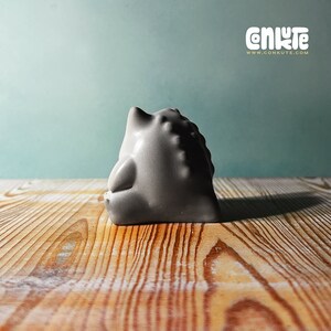 May include: A gray ceramic sculpture of a stylized animal with a rounded shape and textured surface. The sculpture is sitting on a wooden surface.