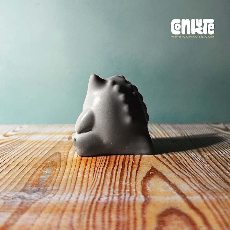 May include: A gray ceramic sculpture of a stylized animal with a rounded shape and textured surface. The sculpture is sitting on a wooden surface.