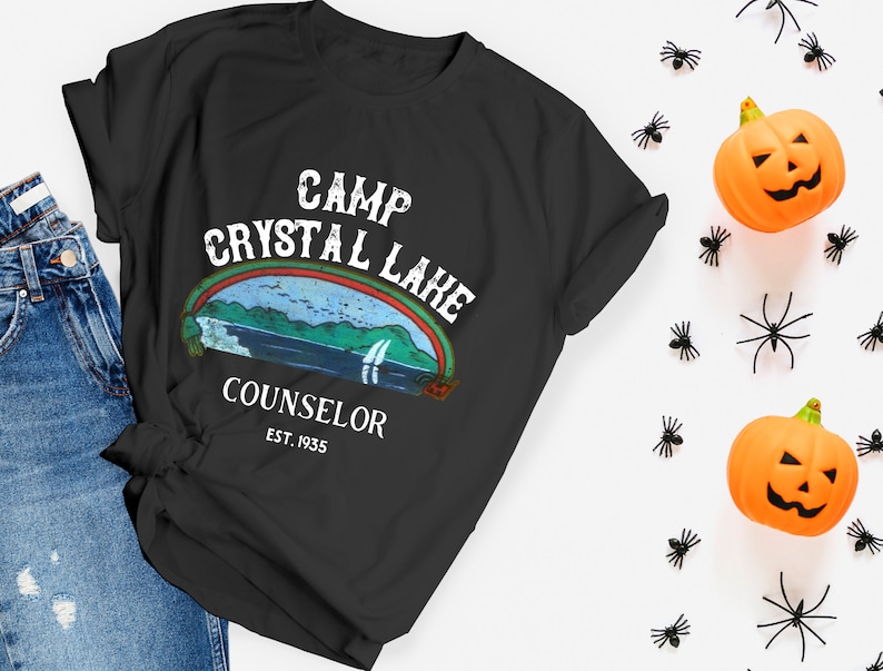 Pu&ograve; includere: Maglietta nera con una grafica in stile vintage che dice "Camp Crystal Lake Counselor Est. 1935". La grafica presenta un lago con un arcobaleno e montagne sullo sfondo.