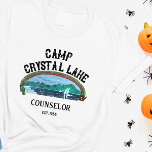 Pu&ograve; includere: Maglietta bianca con una grafica in stile vintage che dice "Camp Crystal Lake Counselor Est. 1935". La grafica presenta un arcobaleno, un lago e alberi.