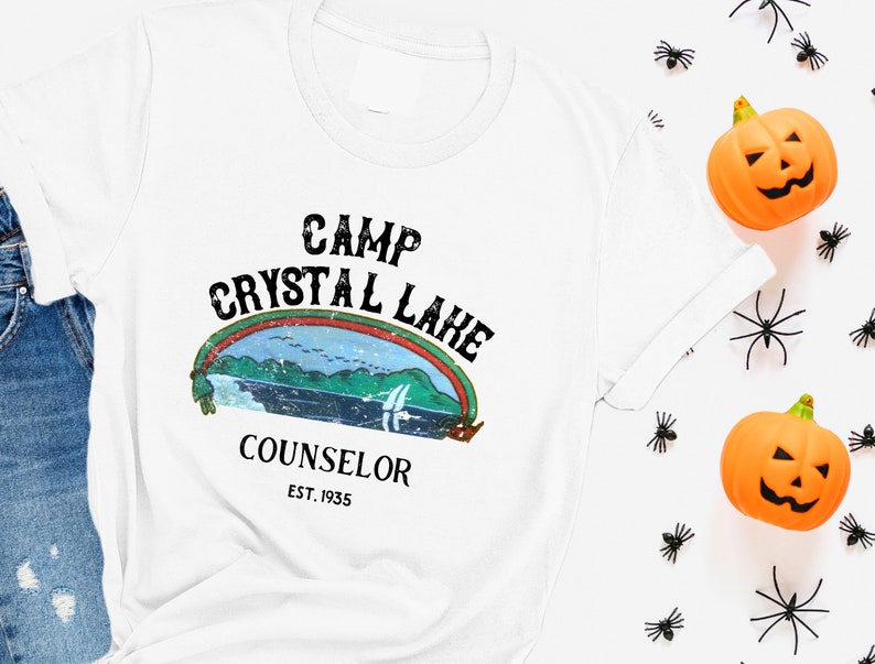 Pu&ograve; includere: Maglietta bianca con una grafica in stile vintage che dice "Camp Crystal Lake Counselor Est. 1935". La grafica presenta un arcobaleno, un lago e alberi.