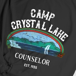 Pu&ograve; includere: T-shirt nero con un design grafico bianco che dice "Camp Crystal Lake Counselor Est. 1935". La grafica presenta un'illustrazione in stile vintage di un lago con montagne e un arcobaleno.