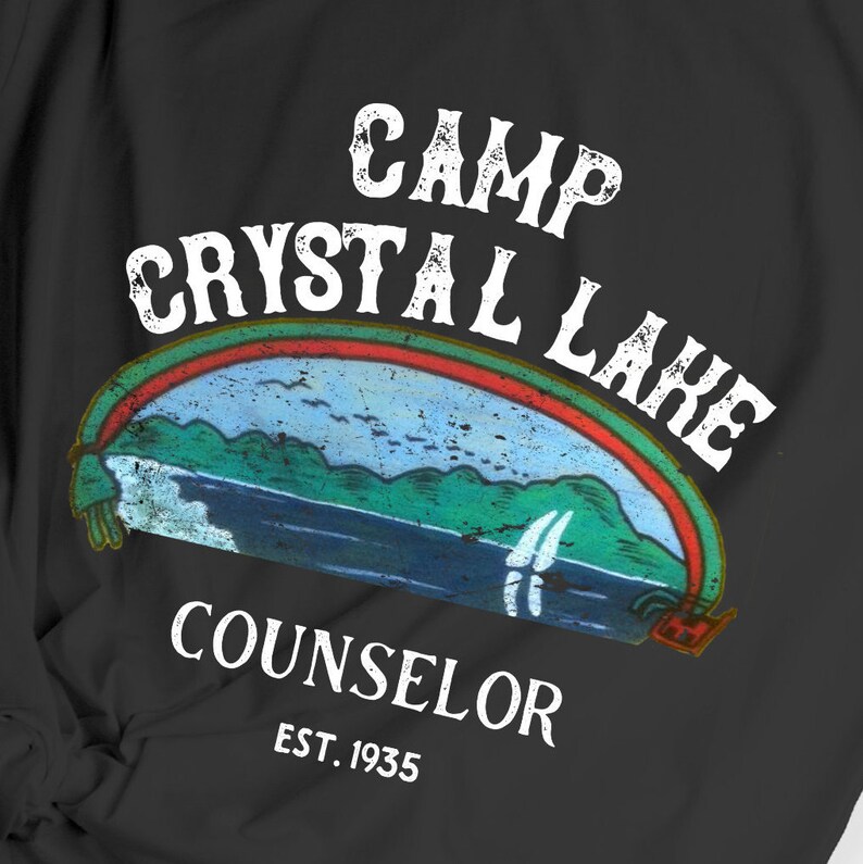 Pu&ograve; includere: T-shirt nero con un design grafico bianco che dice "Camp Crystal Lake Counselor Est. 1935". La grafica presenta un'illustrazione in stile vintage di un lago con montagne e un arcobaleno.
