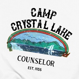 Pu&ograve; includere: Un design grafico in stile vintage con il testo "Camp Crystal Lake Counselor Est. 1935". Il design presenta un arcobaleno sopra una scena di lago con montagne e una barca a vela.