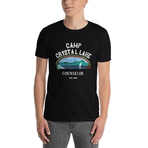 Pu&ograve; includere: Maglietta nera con una grafica vintage di una scena lacustre e il testo "Camp Crystal Lake Counselor Est. 1955".