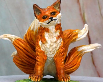 May include: A hand-painted figurine of a kitsune, a Japanese mythical fox with nine tails. The fox is orange and white with a green base.