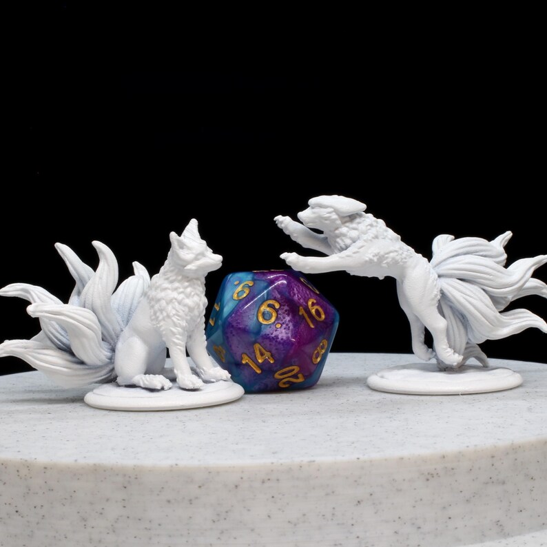 May include: Two white nine-tailed fox figurines positioned on either side of a multi-coloured twenty-sided die. One fox is sitting upright while the other is in a leaping pose. The die features a purple and blue gradient with gold-coloured numbers.