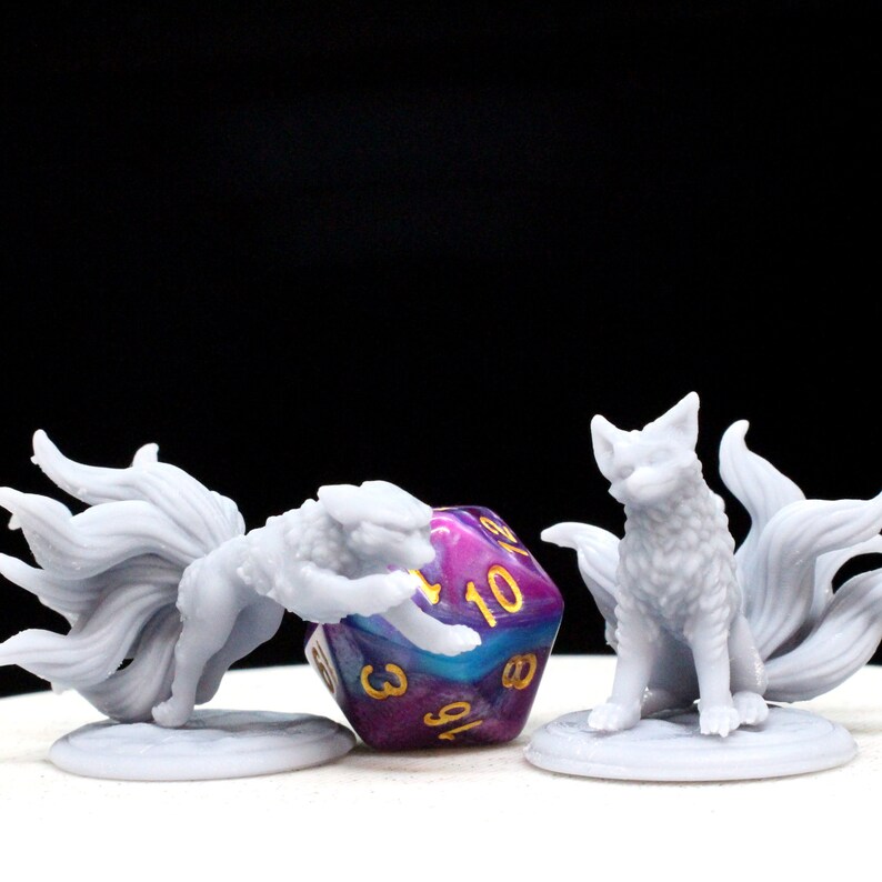 3D printed monster miniatures for tabletop games next to a 20 sided die.  There are 2 figures, both foxes with 9 tails, one leaping forward to attack and one sitting down.