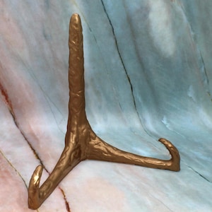 May include: A gold-colored metal display stand with a textured surface and three legs. The stand is designed to hold a plate or other flat object.