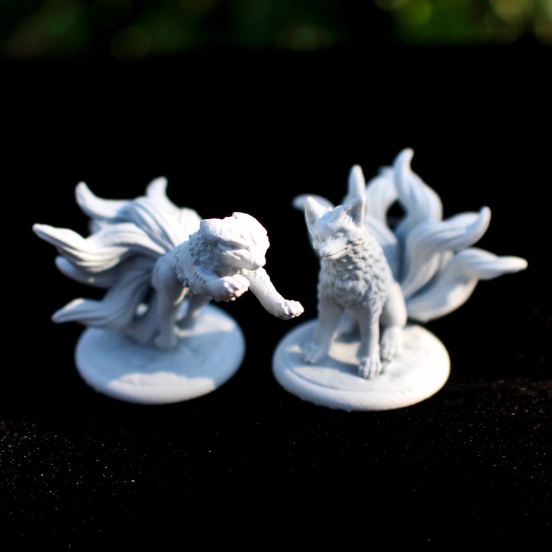 May include: Two unpainted light grey miniature figurines of nine-tailed foxes. One fox is in a pouncing pose with its front paw extended, while the other sits upright. Both figures feature detailed fur textures and multiple tails, mounted on circular bases.