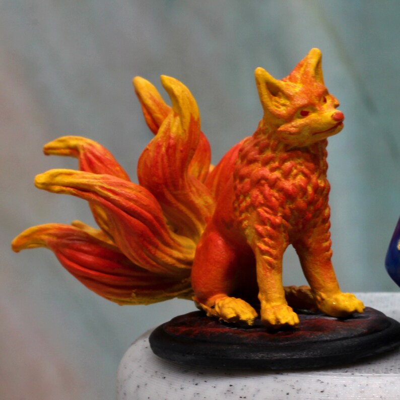 May include: A hand-painted miniature figurine of a nine-tailed fox creature. The fox is coloured in vibrant shades of orange, red, and yellow, featuring a textured fur pattern and multiple flowing tails. It is mounted on a circular black base.