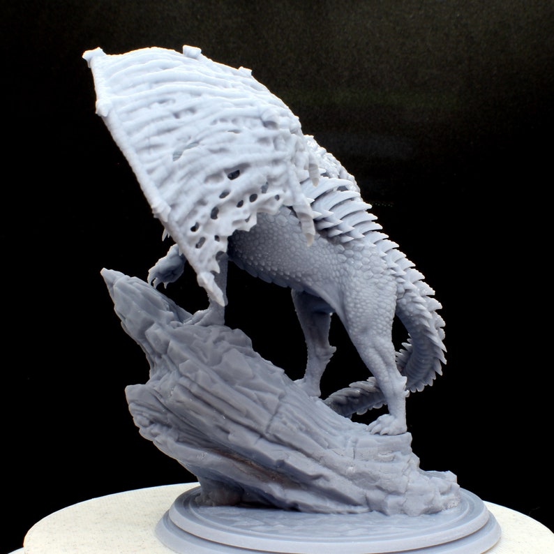 Puede incluir: Un drag&oacute;n en miniatura gris, impreso en 3D, con una envergadura de alas &oacute;seas detallada, posado sobre una base rocosa. El drag&oacute;n est&aacute; en posici&oacute;n con la cabeza girada hacia atr&aacute;s, mirando por encima del hombro.