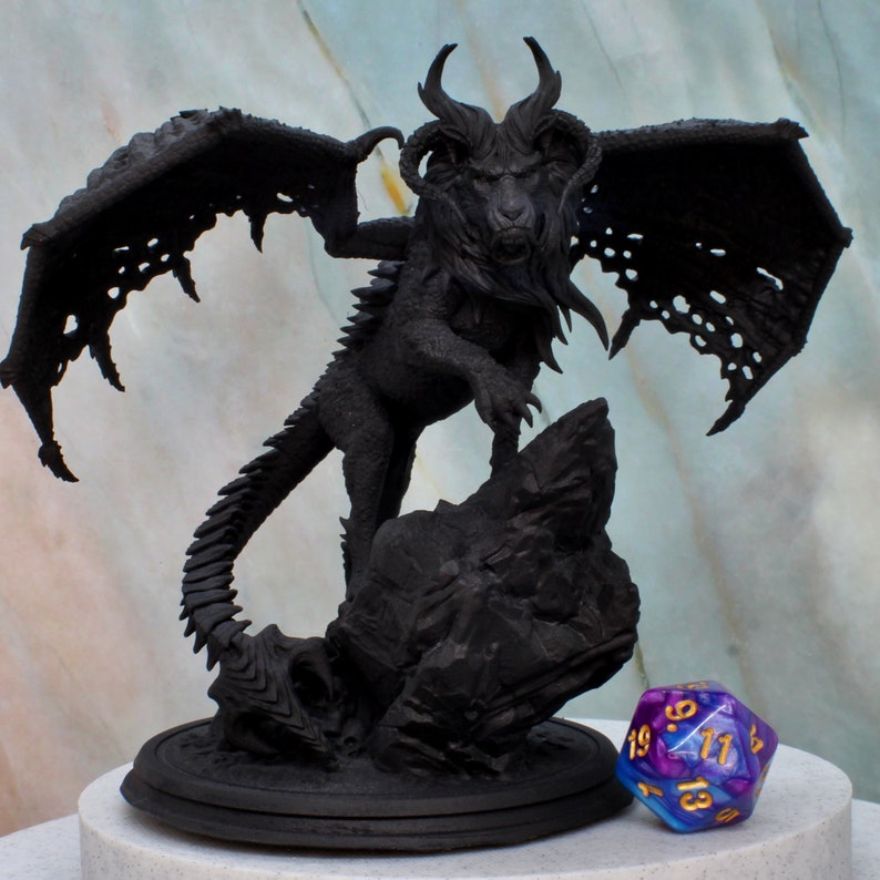 Puede incluir: Una estatua de drag&oacute;n negro con las alas extendidas, de pie sobre una base rocosa. El drag&oacute;n tiene una expresi&oacute;n feroz y garras afiladas. Un dado morado y azul con n&uacute;meros dorados est&aacute; en primer plano.