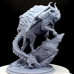 Puede incluir: Una miniatura gris, impresa en 3D, de una criatura de le&oacute;n alado de pie sobre una base de roca. La criatura tiene una cola larga y puntiaguda y una melena de pelo.