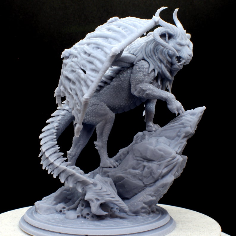 Puede incluir: Una miniatura gris, impresa en 3D, de una criatura de le&oacute;n alado de pie sobre una base de roca. La criatura tiene una cola larga y puntiaguda y una melena de pelo.