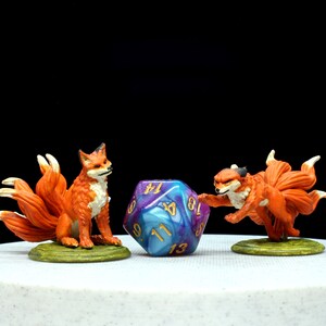 May include: Two miniature orange foxes with multiple tails, each standing on a circular green base, positioned on either side of a large, twenty-sided die featuring a swirling blue and purple marbled pattern with gold-coloured numbers.