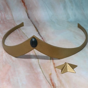 May include: A gold-colored crown with a dark green oval gem in the center. A small, gold star-shaped accessory is next to the crown. The crown is a simple band with a slight upward curve at the center.