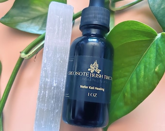 Può includere: Una bottiglia di vetro scuro con un contagocce, etichettata "CREOSOTE BUSH TINCTURE" e "Nefer Kali Healing", accanto a un cristallo di selenite bianco. La bottiglia contiene un liquido scuro. Foglie verdi e viti sullo sfondo.