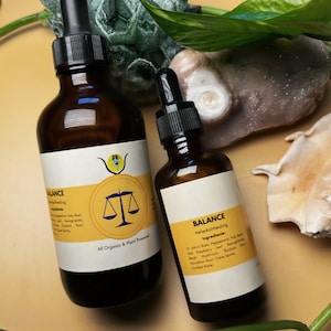 May include: Two amber glass dropper bottles with black caps, labelled "BALANCE." The labels feature a yellow circle with a scale illustration and the text "All Organic & Plant Powered." Ingredients are listed. The bottles are on a light brown surface.