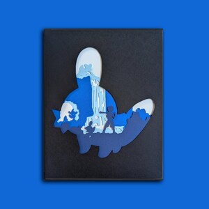 May include: A layered paper art piece in a black frame featuring a silhouette of a creature. The cutout reveals a blue-toned scene with a waterfall, rocky terrain, and a small figure standing in the center, rendered in shades of light and dark blue.