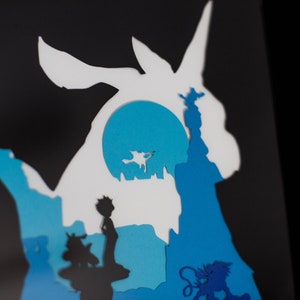 May include: Paper cut-out art depicting a blue and white silhouette of a Pokemon character, with a blue moon in the background. The character is surrounded by other smaller cut-out figures, including a person and a small animal.