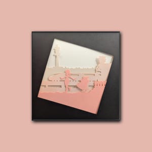 May include: A framed paper art piece featuring a layered design of a park scene. The artwork depicts a girl, a dog, and a building, all in a soft pink hue, set against a white and light gray background. The frame is black.