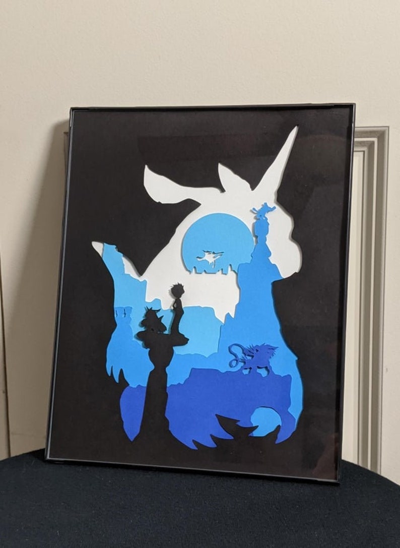 May include: A framed paper art piece depicting a blue and white silhouette of a mythical creature with a unicorn horn. The creature is surrounded by a blue and white landscape with a small figure standing on a tower.