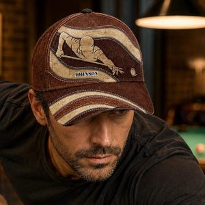 May include: A brown baseball cap with a billiards design. The cap features a tan illustration of a person playing pool, with the name "JOHNSON" printed on the front. The cap has a curved brim and a dark brown crown.