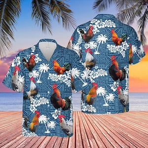 Puede incluir: Una camisa de manga corta con un diseño de gallo y flores sobre un fondo azul. El patrón incluye flores blancas y palmeras, dando una sensación tropical. La camisa se muestra sobre una superficie de madera con un fondo de playa y puesta de sol.