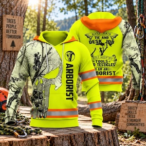 Pode incluir: Uma camisola com capuz amarelo néon e forro laranja, com um gráfico de um arborista a trepar, uma silhueta de caveira e o texto "KEEP YOUR TIE AND DESK JOB WILL KEEP MY TOOLS AND TESTICLES I AM AN ARBORIST" com faixas refletoras.