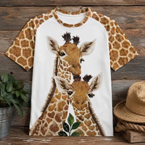 May include: A short-sleeve t-shirt featuring a white centre panel with a graphic illustration of two giraffes and green leaves. The sleeves and neckline are patterned with a brown and tan giraffe print, creating a cohesive animal-themed design.