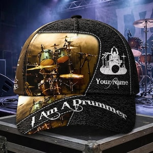 May include: A black baseball cap with a textured leather-like finish featuring a printed graphic of a drum kit on stage. The cap includes the text "Your Name" and "I Am A Drummer" in white lettering, with a stylised drum set icon above the name.