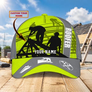 May include: A neon yellow and gray baseball cap featuring a graphic of two roofers working on a wooden roof frame. The cap includes the text ROOFER and YOUR NAME, along with a small icon of a roof and a circular tool emblem on the brim.