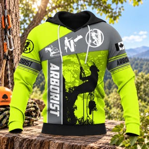 Pode incluir: Um moletom com capuz amarelo neon e cinza apresentando a silhueta preta de um arborista subindo em uma árvore. O design inclui a palavra ARBORIST em texto branco, vários gráficos circulares de trabalhadores florestais e uma pequena bandeira canadense.