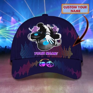 Puede incluir: Una gorra de béisbol azul oscuro con un gráfico de un DJ mezclando en tocadiscos, auriculares y un patrón de ondas sonoras de colores. El texto "YOUR NAME" está impreso en morado y un recuadro arriba dice "CUSTOM YOUR NAME".