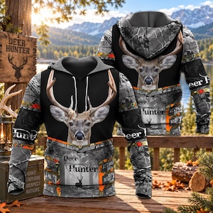May include: A long-sleeve hooded sweatshirt featuring a realistic deer head graphic on the front and back. The design incorporates a black center panel, grey camouflage patterns, and orange accents. The text "Deer Hunter" is printed on the garment.