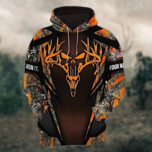 May include: A long-sleeve hooded sweatshirt featuring an orange and black camouflage pattern. The front displays a large orange deer skull graphic with antlers. The text "HUNTER" and "YOUR NAME" appear on the sleeves, with a small US flag patch.