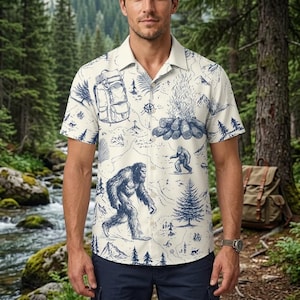 May include: A short-sleeve button-down shirt in off-white featuring a blue ink-style print of a map with a backpack, a campfire, pine trees, mountains, and a large hairy creature resembling Bigfoot walking across the landscape.