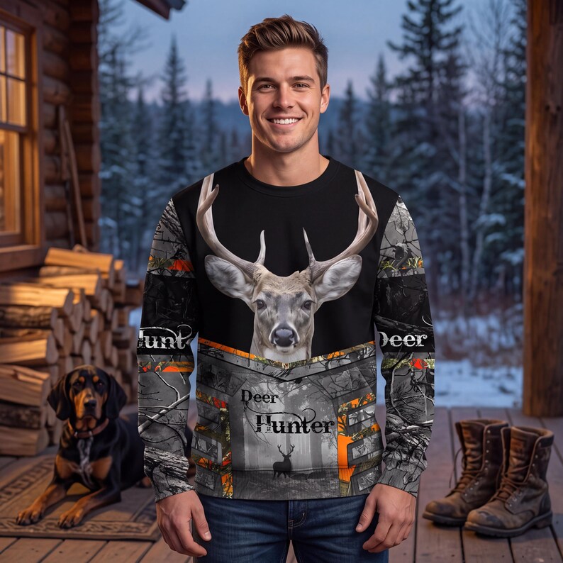 May include: A long-sleeve sweatshirt featuring a large deer head graphic on the chest, a camouflage pattern in gray and orange, and the text "Deer Hunter" printed on the front. The garment has a crew neckline and a black upper panel.