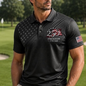 Puede incluir: Un polo negro con un patr&oacute;n sutil de estrellas en el pecho derecho y un gr&aacute;fico en el pecho izquierdo que dice 250 FREEDOM 1776-2026 con un dise&ntilde;o de bandera ondeante. Un peque&ntilde;o parche de la bandera estadounidense es visible en la manga izquierda.