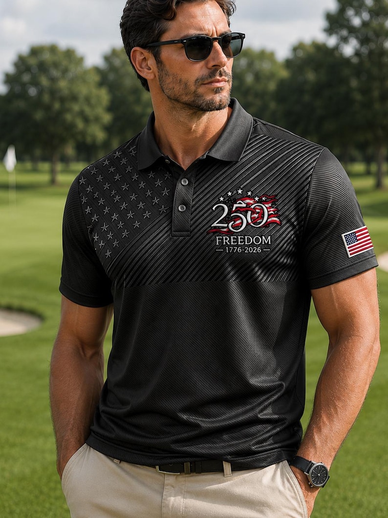 Puede incluir: Un polo negro con un patr&oacute;n sutil de estrellas en el pecho derecho y un gr&aacute;fico en el pecho izquierdo que dice 250 FREEDOM 1776-2026 con un dise&ntilde;o de bandera ondeante. Un peque&ntilde;o parche de la bandera estadounidense es visible en la manga izquierda.