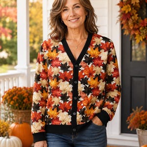May include: A long-sleeve button-down cardigan featuring a vibrant autumn leaf pattern in shades of red, orange, yellow, and black. The garment has a solid black V-neck collar, black cuffs, and a black hem, paired with blue denim jeans.