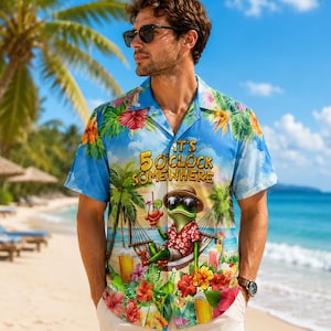Könnte beinhalten: Hellblaues Hawaiihemd mit tropischem Motiv. Es zeigt einen Frosch mit Sonnenbrille, der sich in einer Hängematte mit Cocktail entspannt. Der Text "It's 5 O'Clock Somewhere" ist von Palmen und Blumen umgeben.