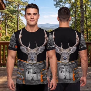 May include: A black short-sleeve t-shirt featuring a large deer head graphic on the chest, with the text "Deer Hunter" printed below. The lower portion and sleeves display a gray forest camouflage pattern with orange accents and silhouettes.