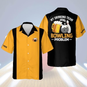Puede incluir: Una camisa de bolos de manga corta con un diseño de bloques de color amarillo y negro. La camisa presenta un gráfico de una jarra de cerveza, una bola de bolos y un pin, con el texto "MY DRINKING TEAM HAS A BOWLING PROBLEM". La palabra "HERE" está impresa en la manga izquierda.