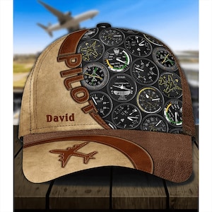 May include: A brown and tan baseball cap with a pilot theme. The cap features a variety of aircraft gauges and the word "Pilot" in a brown, stitched font. The name "David" is embroidered on the front of the cap.