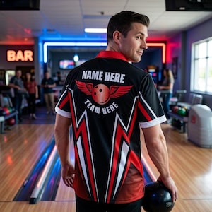 Peut inclure: T-shirt de bowling noir avec des accents rouges et blancs. Le t-shirt présente un motif de boule de bowling ailée et le texte "NAME HERE" et "TEAM HERE". La personne tient une boule de bowling noire.