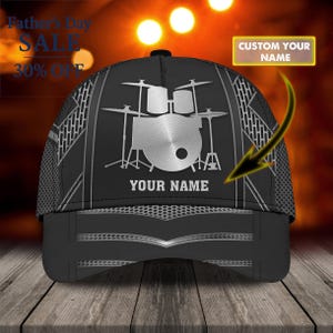 Op de afbeelding: Zwarte baseballcap met een zilveren drumstelontwerp en de tekst "YOUR NAME". De cap heeft een mesh achterkant en een gebogen klep. De afbeelding bevat ook de tekst "Father's Day SALE 30% OFF" en "CUSTOM YOUR NAME".